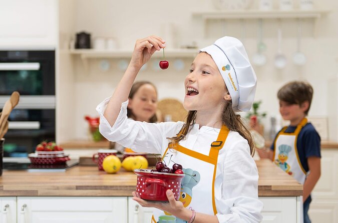 Mädchen genießt Kirsche – fröhlicher Moment in der Kinderkochschule Ein Mädchen im Vordergrund isst eine Kirsche, im Hintergrund stehen ein Junge und ein Mädchen in der Küche.