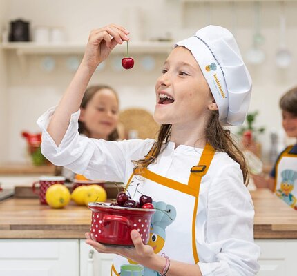 Mädchen genießt Kirsche – fröhlicher Moment in der Kinderkochschule Ein Mädchen im Vordergrund isst eine Kirsche, im Hintergrund stehen ein Junge und ein Mädchen in der Küche.