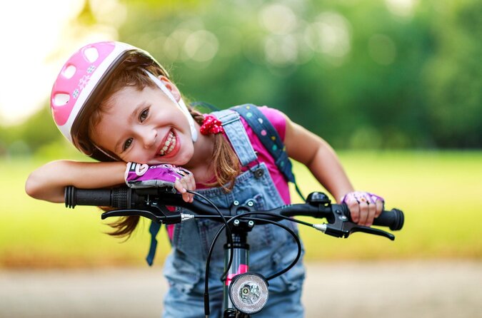 Strahlendes Lächeln im Sattel – so schön kann Familienurlaub mit dem Rad sein. Lächelndes Mädchen mit Fahrradhelm sitzt auf einem Kinderfahrrad bei sonnigem Wetter.